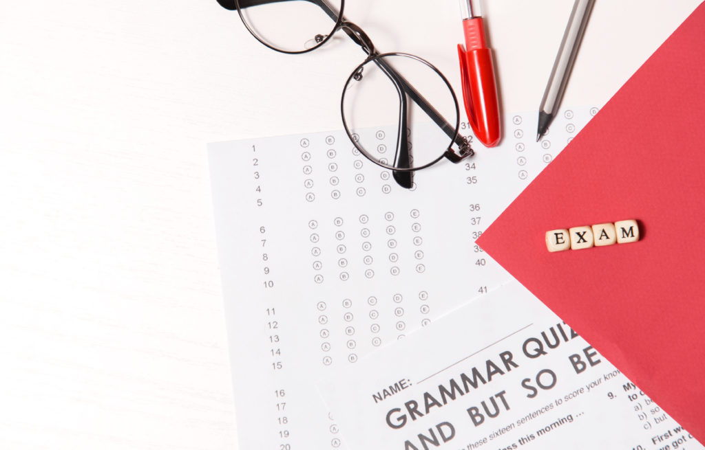 Glasses, pencil, and pen sit on top of a multiple-choice test, four block letters spell the word “exam.”