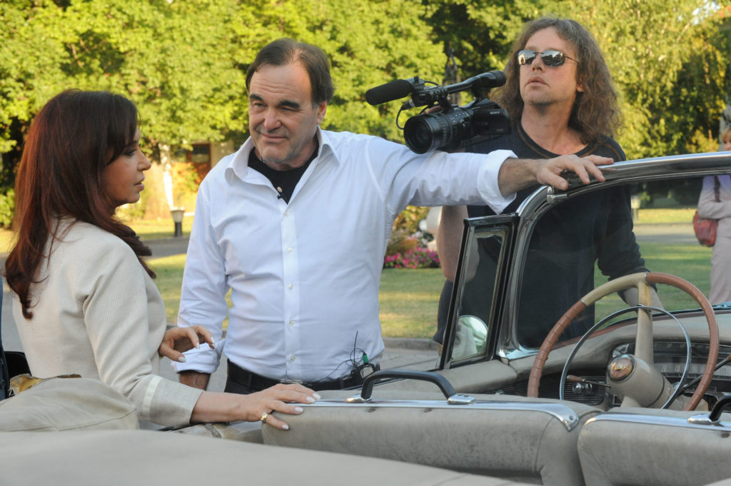 Photo of Oliver Stone standing with a movie camera near a vintage car